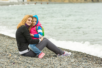 Image showing Mom and daughter in the arms sit on the beach on a cool day and watching the surf