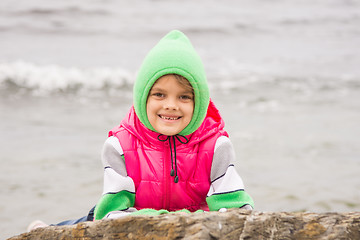 Image showing Warmly dressed girl with a smile peeking out from behind a rock against the backdrop of the sea