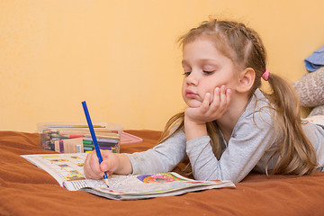 Image showing Little girl studying a magazine with a pencil in his hand lying on his stomach and his head in his second hand