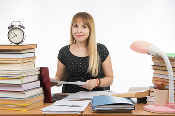 Image showing The girl behind the desk littered with books with a smile, holding a paper