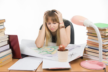 Image showing A girl student at a table with a pile of books, drawings and projects sits holding his head in his hands