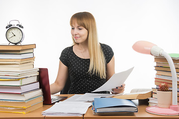 Image showing Joyful girl behind a desk littered with books, papers in hand looking in laptop