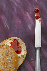 Image showing Halved wholemeal roll with butter and red jam and knife with jam