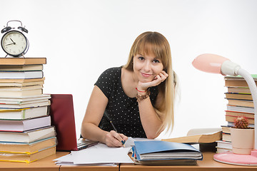 Image showing  She is engaged at the table cluttered with books in the library with a smile looks in the frame