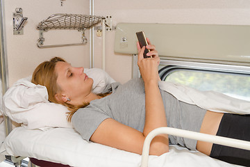 Image showing The girl on the train lying on his back on the top shelf in the side car with reserved seats and looking at the phone