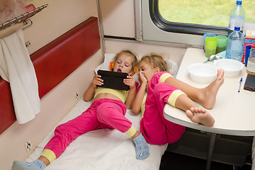 Image showing Two little girls lie on the train with his feet on the lower second-class place car in the same pajamas and looking at tablet