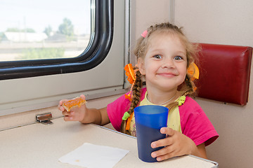 Image showing Little girl with a happy face drink tea with a sandwich on the train at the table on outboard second-class carriage