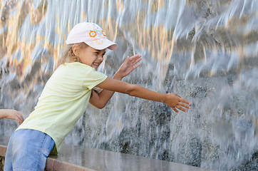 Image showing Cheerful six year old girl in summer clothes hand trying to get the water artificial waterfall