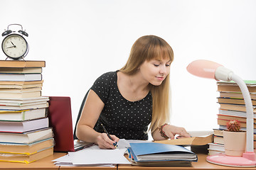 Image showing Pretty female student at a table littered with books in the library writing job