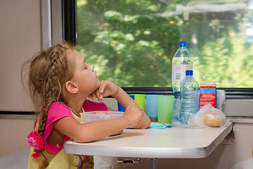 Image showing A six-year girl in a train sitting at a table with food on a lower place in the second-class compartment of the car and looks thoughtfully out the window