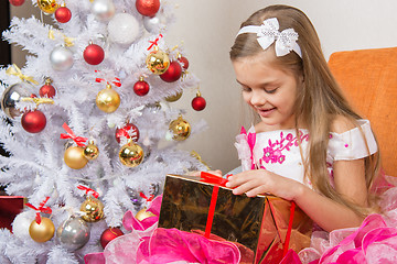 Image showing Seven-year girl in a beautiful dress is considering a gift sitting on the couch at Christmas tree