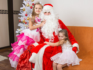 Image showing Two girls in beautiful dresses hug Santa Claus sitting on a couch