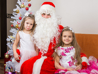 Image showing Two girls in beautiful dresses hug Santa Claus sitting on the couch, one of them a little displeased