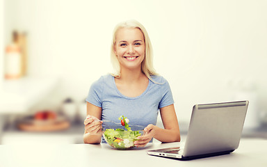 Image showing smiling woman eating salad with laptop on kitchen