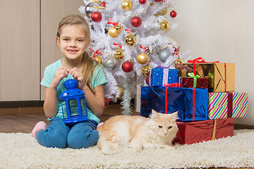 Image showing Seven-year girl sits with a cat under the Christmas tree with gifts and smiling happily