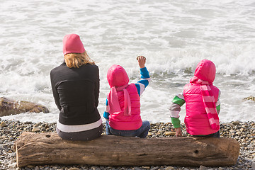 Image showing Two girls and woman sitting on a log on the beach and throwing stones into the water