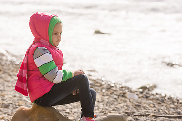 Image showing Thoughtful girl sitting on a rock on the stony beach of the sea and looks into the distance