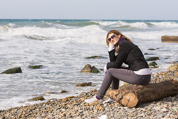 Image showing Happy girl with glasses sitting on a log on a rocky beach ocean