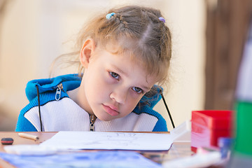 Image showing The sad girl sits at a table in the courtyard of the house and does not want to draw