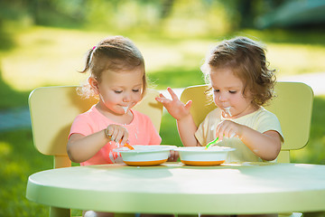 Image showing Two little girls sitting at a table and eating together against green lawn