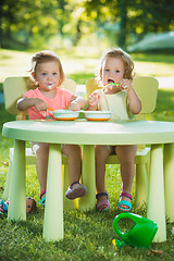 Image showing Two little girls sitting at a table and eating together against green lawn