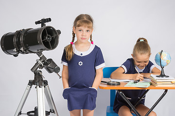 Image showing A girl stands at a telescope with his hands in his pockets, another girl having fun sitting at the table and writes