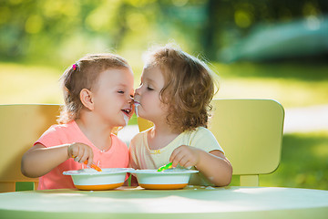 Image showing Two little girls sitting at a table and eating together against green lawn