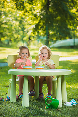 Image showing Two little girls sitting at a table and eating together against green lawn