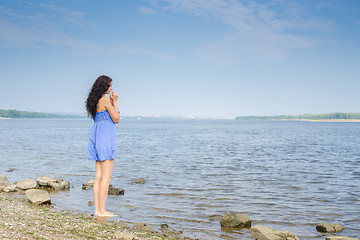 Image showing Sad young brunette woman in a blue summer dress standing on the river bank