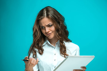 Image showing The displeased young business woman with pen and tablet for notes on blue background
