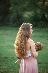 Image showing Young beautiful mother hugging her little toddler son against green grass. Happy woman with her baby boy on a summer sunny day. Family walking on the meadow.