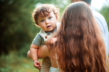 Image showing Young beautiful mother hugging her little toddler son against green grass. Happy woman with her baby boy on a summer sunny day. Family walking on the meadow.