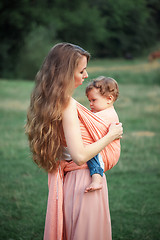 Image showing Young beautiful mother hugging her little toddler son against green grass. Happy woman with her baby boy on a summer sunny day. Family walking on the meadow.