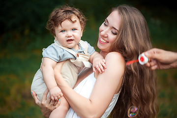 Image showing Young beautiful mother hugging her little toddler son against green grass. Happy woman with her baby boy on a summer sunny day. Family walking on the meadow.