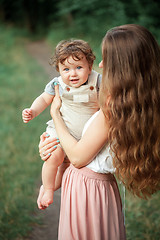 Image showing Young beautiful mother hugging her little toddler son against green grass. Happy woman with her baby boy on a summer sunny day. Family walking on the meadow.