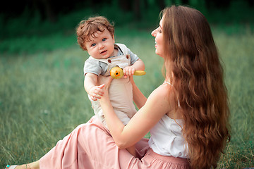 Image showing Young beautiful mother sitting with her little son against green grass. Happy woman with her baby boy on a summer sunny day. Family walking on the meadow.