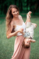 Image showing Young beautiful mother hugging her little toddler son against green grass. Happy woman with her baby boy on a summer sunny day. Family walking on the meadow.