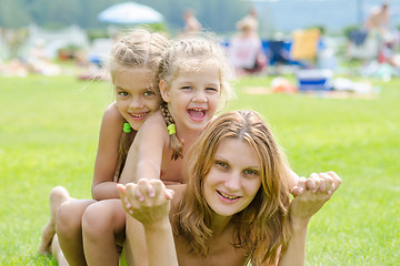 Image showing Two daughters are sitting on the mother\'s back, hot summer day relaxing on the green lawn beautiful beaches