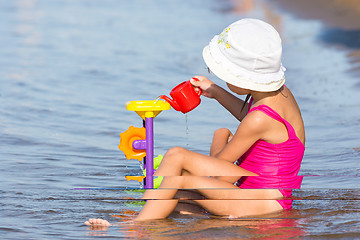 Image showing Five-year girl in a pink bathing suit sitting in on the river bank and plays in the sand toys