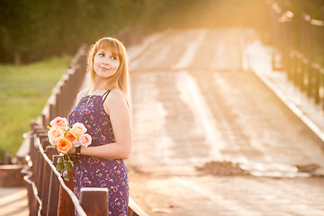 Image showing The charming young girl standing on a rustic bridge at dawn sun with a bouquet of roses