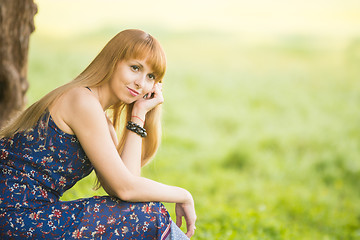 Image showing Beautiful young girl sitting against a tree on the background of blurred green grass and looks into the distance
