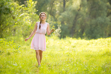 Image showing Young beautiful woman walking on a green meadow with a bouquet of wildflowers in a light summer dress