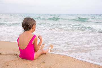 Image showing The girl in the pink bathing suit sitting on the beach, eating a wafer and looks into the distance