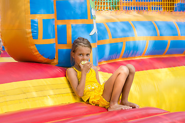 Image showing Girl eats an apple sitting on the edge of the inflatable trampoline in amusement park