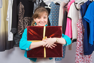 Image showing Happy young girl standing with big red gift on background of clothes on hangers