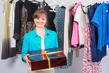 Image showing Happy young girl standing with a red big present, the background hang things on hangers