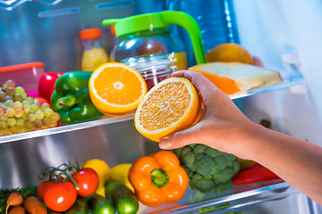 Image showing Woman takes the orange from the open refrigerator.