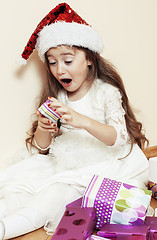 Image showing little cute girl in santas red hat waiting for Christmas gifts. smiling adorable kid. White new dress home interior 