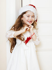 Image showing little cute girl in santas red hat waiting for Christmas gifts. smiling adorable kid. White new dress home interior