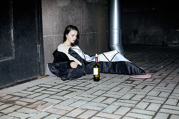 Image showing young poor ttenage girl sitting at dirty wall on floor with bottle of vine, poor refugee alcoholic, hopeless homeless woman in depression, real junky concept